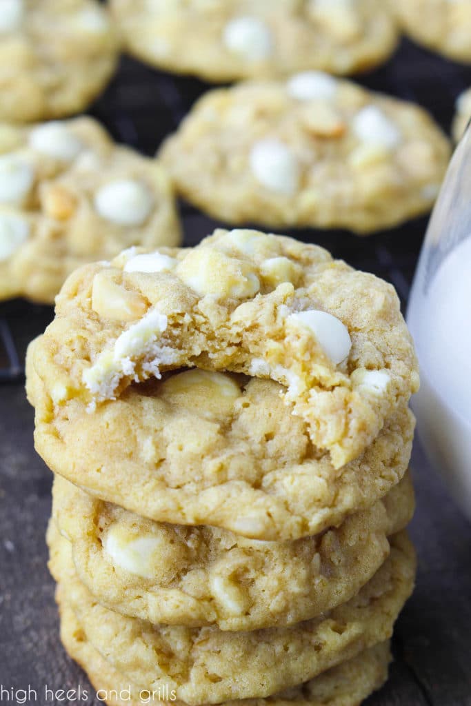 Stack of White Chocolate Chip Macadamia Nut cookies with a bite taken out of the top cookie. Paired with a glass of milk and a cooling rack of cookies in the background.