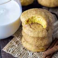 Stack of pumpkin snickerdoodles recipe with a bite taken out of the top cookie. Glass of milk on one side and cinnamon sticks on the other side.