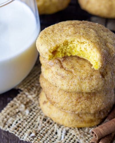 Stack of pumpkin snickerdoodles recipe with a bite taken out of the top cookie. Glass of milk on one side and cinnamon sticks on the other side.