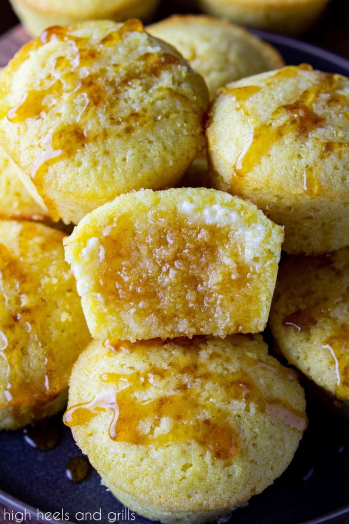 A pile of sweet cornbread muffins with honey drizzled on the tops. The middle one split in half, butter slathered on it, and honey soaking into it.