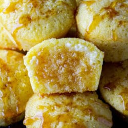 A pile of sweet cornbread muffins with honey drizzled on the tops. The middle one split in half, butter slathered on it, and honey soaking into it.