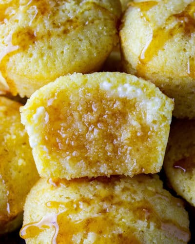 A pile of sweet cornbread muffins with honey drizzled on the tops. The middle one split in half, butter slathered on it, and honey soaking into it.