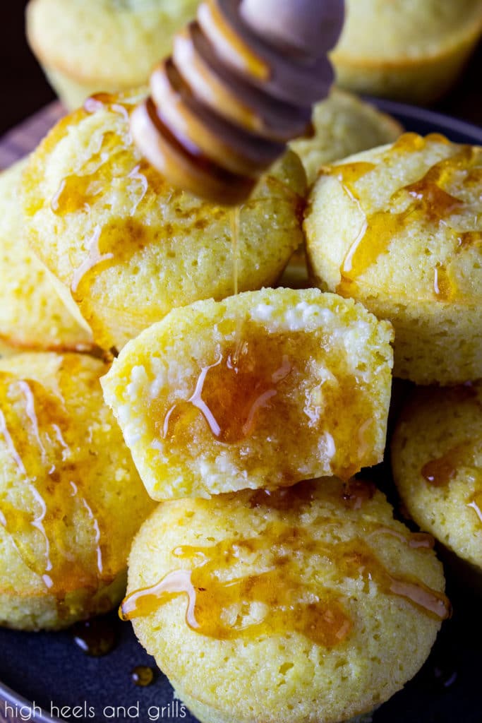 A pile of sweet cornbread muffins with honey drizzled on the tops. The middle one split in half, butter slathered on it, and honey being drizzled from a honey dipper.